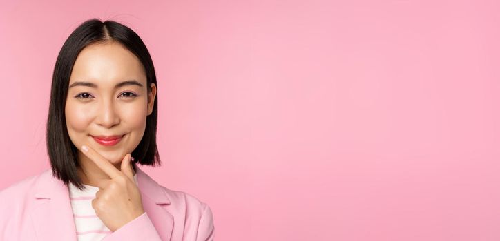 Close up portrait of smiling asian working lady in suit, businesswoman looking thoughtful, thinking or deciding smth, standing over pink background