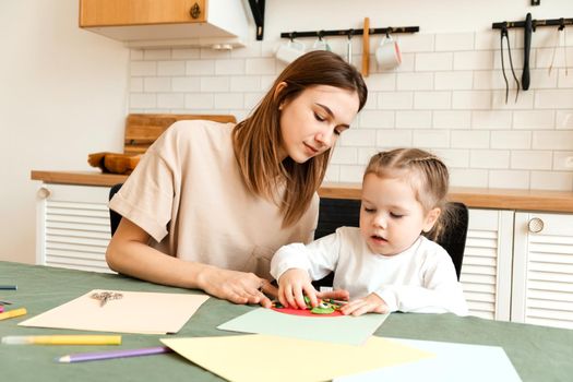 Young mother helping cute daughter with art and craft project in home interior. Babysitter teaches child to do crafts.