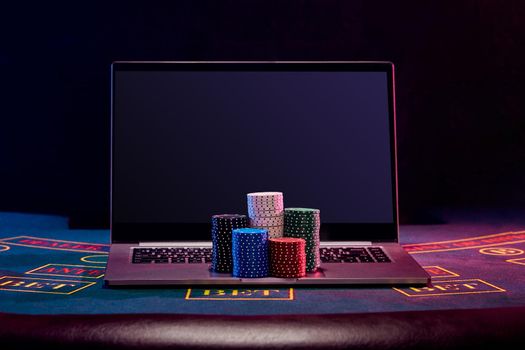 Chips piles are on a laptop standing on blue cover of playing table. Black background. Gambling entertainment, poker, casino concept. Close-up.