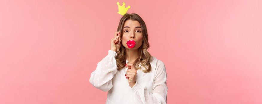 Spring, happiness and celebration concept. Close-up portrait of funny and cute, silly blond girl in white dress, holding big lips mask and crown, partying and having fun, pink background