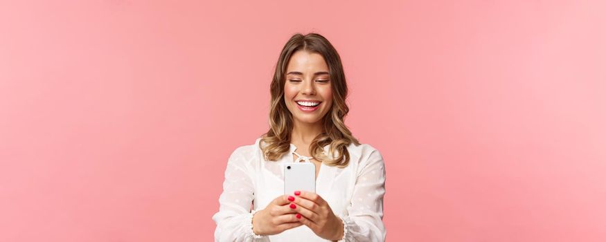 Close-up portrait of happy charming caucasian woman with short fair hair, white dress, record video of concert on mobile phone, taking photo using smartphone, standing pink background