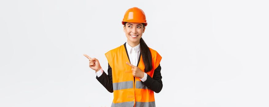 Building, construction and industrial concept. Smiling confident asian female chief engineer in safety helmet and reflective clothing pointing fingers upper left corner and looking satisfied