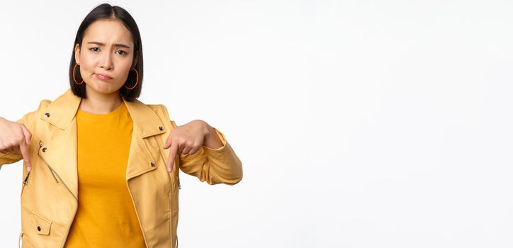 Portrait of disappointed asian woman pointing fingers down, grimacing and showing smth bad, standing over white background