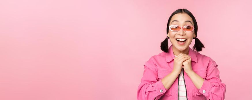 Portrait of excited japanese girl in sunglasses, celebrating, achieve goal, gasping amazed and smiling, standing over pink background