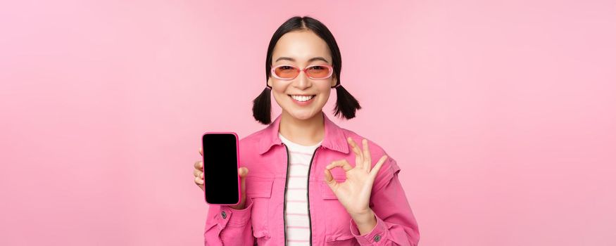 Enthusiastic young asian woman showing okay, ok sign, smiling pleased, mobile phone screen, smartphone application, standing over pink background