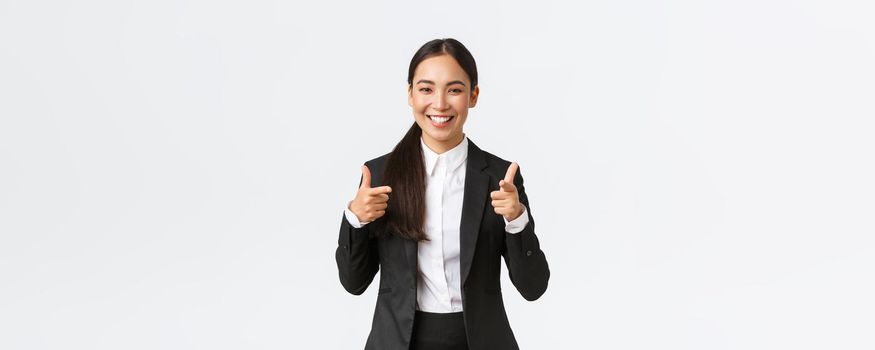 Confident smiling asian saleswoman in black suit showing thumbs-up, guarantee quality of product or providing best service. Sales manager trying sell something to client, white background
