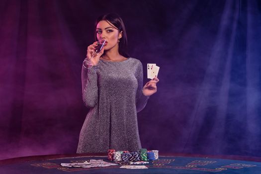 Girl playing poker at casino, holding two aces, drinking champagne. Posing at table with chips, money on it. Black, smoke background. Close-up.