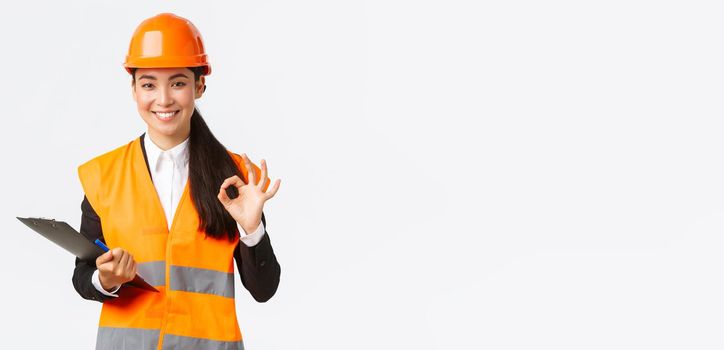 Satisfied smiling asian female architect, construction engineer in helmet and reflective jacket, showing okay gesture, approve and give permission for builing works, finish inspection