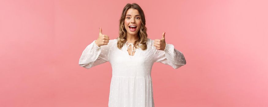 Portrait of feminine blond woman in white dress, show thumbs-up in approval, say yes, approve or agree, satisfied and recommend product, standing pink background in white dress