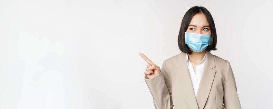 Enthusiastic businesswoman pointing fingers left, wearing medical face mask from covid-19 pandemic, standing over white background