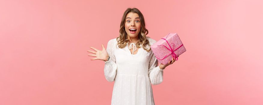 Holidays, celebration and women concept. Portrait of surprised upbeat charismatic blond girl in white dress, spread hands sideways and gasping amazed, receive surprise gift, hold pink box.