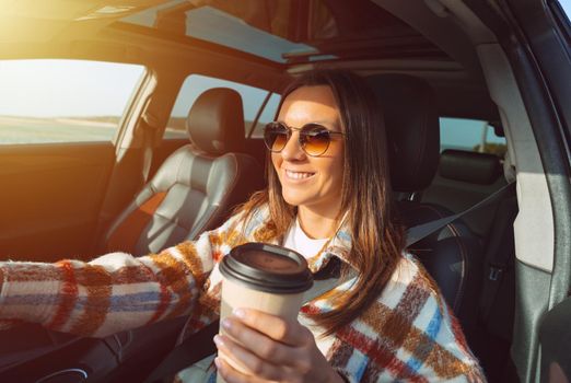 Happy smiling beautiful woman holding coffee cup and driving a car on a sunny day
