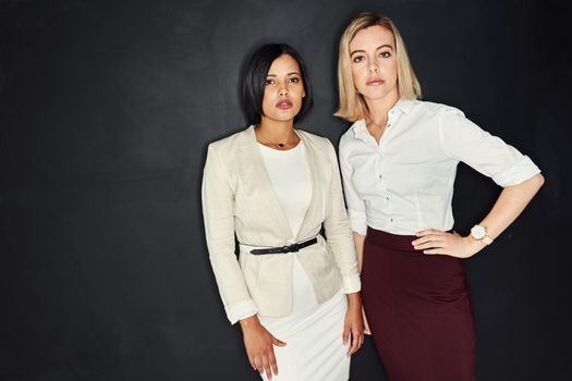 Youve gotta look the part. Portrait of two young businesswomen standing together against a dark background.