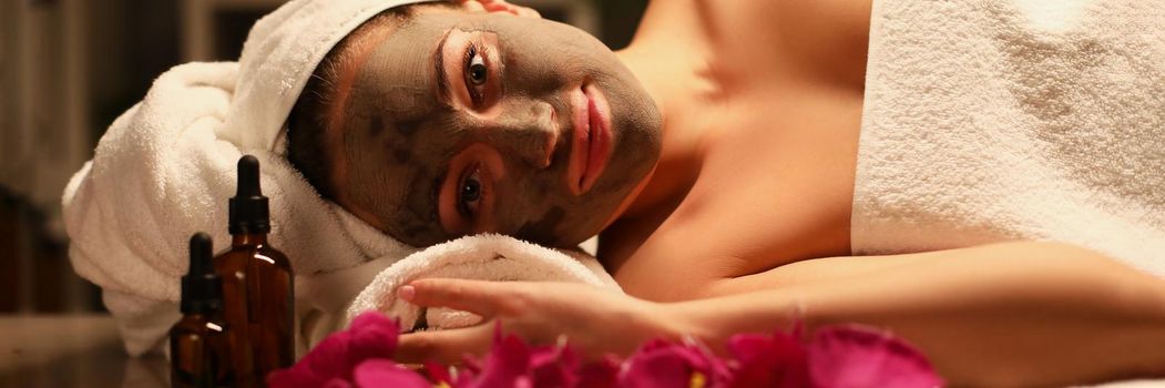 Portrait of smiling relaxed woman in spa centre with clay face mask, luxury spa salon. Rose petals, oil and calm atmosphere. Beauty salon, wellness, body care, chilling concept
