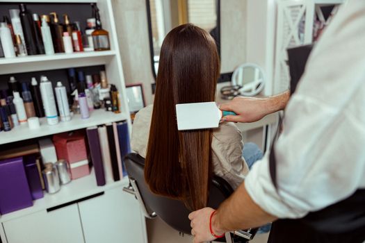 Hairstylist brushing long and sleek brown hair of female client at beauty salon. Hair care