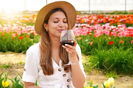 Beautiful woman smells precious red wine with closed eyes in garden with blossomed flowers on background