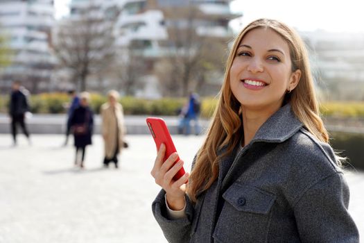 Portrait of smiling woman wearing gray jacket holding smartphone in pedestrian street