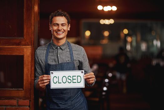 Sorry, come back another time. Portrait of a cheerful young business owner holding up a sign saying closed while standing under a doorway at a beer brewery during the day.