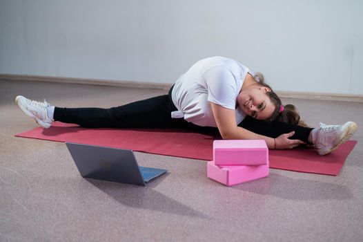 Young caucasian fat woman doing bends on a sports mat and watching a training video on a laptop. A chubby girl stretches the split remotely using video communication