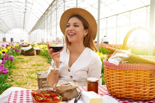 Picnic inside flowers greenhouse with zero kilometer products. Cheerful woman raising a wine glass looking to the side in farmhouse of Tuscany, Italy.