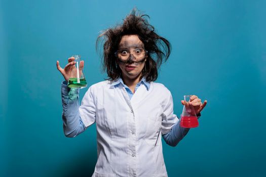 Crazy looking chemist holding Erlenmeyer glass jars filled with unknown chemical compounds while on blue background.