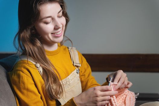Young happy caucasian woman smiling sitting on sofa and crocheting wool product. Women's hobby production of clothes from wool