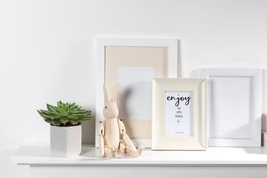 Echeveria in a geometric vase, photo frames and a wooden hare on a chest of drawers against a white wall. Scandinavian style. Ready layout.