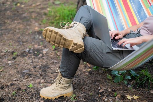Caucasian woman working on laptop while sitting in a hammock in the forest. Girl uses a wireless computer on a hike.