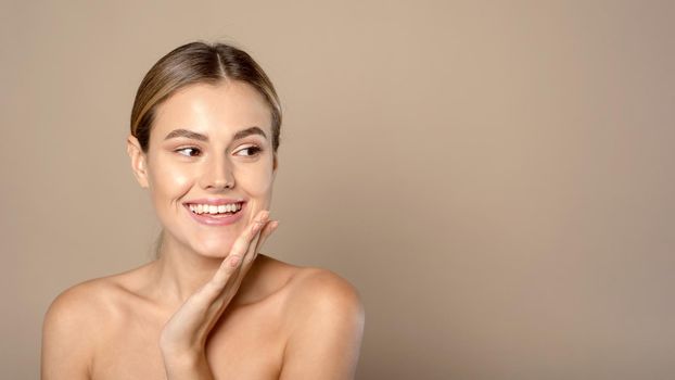 Skin care. Web-banner. Beautiful smiling Caucasian female model with natural makeup touching glowing hydrated skin on beige background