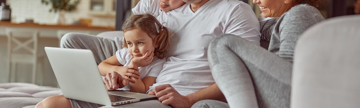 Jolly parents are cuddling with daughter and son while spending time with laptop in living room