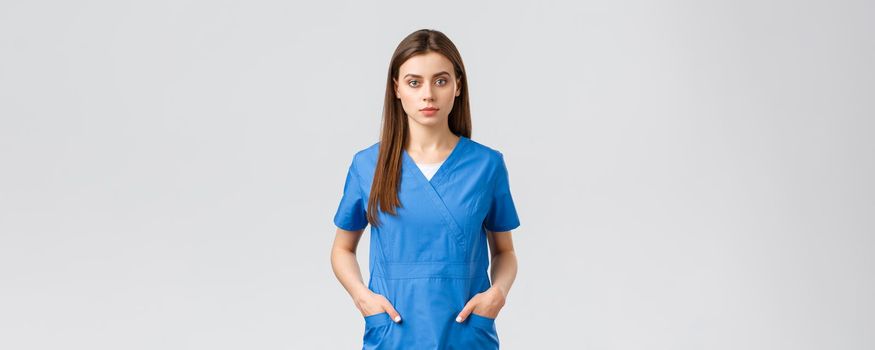 Healthcare workers, prevent virus, insurance and medicine concept. Serious-looking, confident young medical worker, nurse or doctor in blue scrubs hold hands in pockets and looking camera