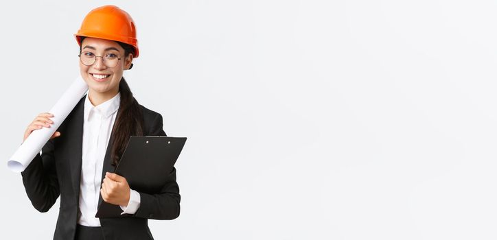 Successful asian female architect in business suit and helmet holding blueprints and clipboard with notes, inspector looking at construction works, smiling at camera, white background