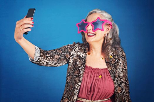 Always capture the fun moments in life. Cropped hot of a funky and cheerful senior woman taking selfies against a blue background.