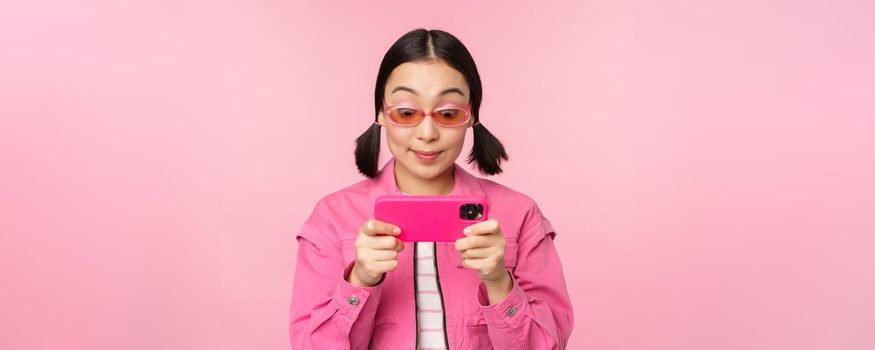 Portrait of happy asian girl playing on smartphone, watching videos on mobile phone app, standing over pink background