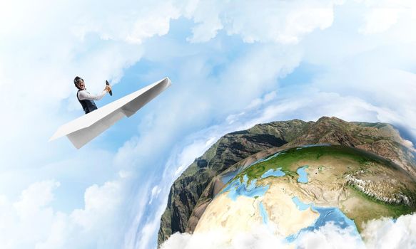 Pilot in leather helmet and goggles driving paper plane on background of blue cloudy sky. Traveling around the world. Funny man flying in small paper airplane. Earth horizon with high mountain range