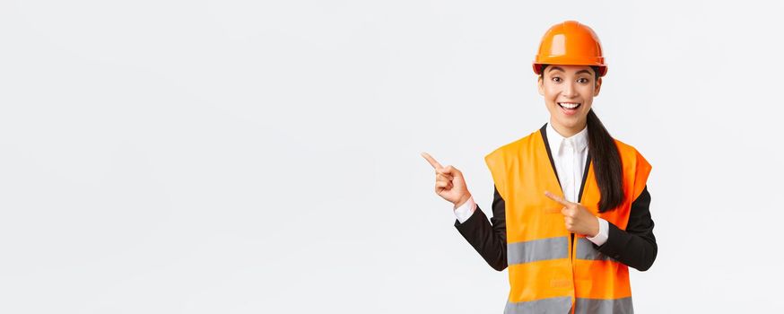 Building, construction and industrial concept. Smiling female engineer in helmet and reflective clothing showing way, pointing upper left corner, talking about constructing project, white background