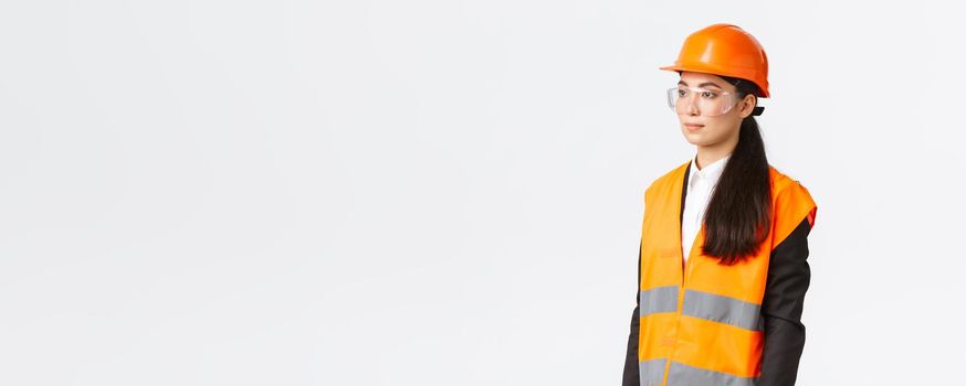 Profile of serious asian female businesswoman inspecting construction area, chief engineer looking left, wearing safety helmet and reflective clothing, standing white background