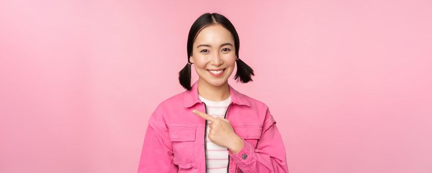 Portrait of beautiful smiling asian girl, pointing finger left, showing advertisement, banner promo, standing over pink background