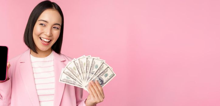 Successful young asian businesswoman showing money, cash dollars and smartphone screen, smiling pleased, pink background