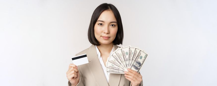 Close up of asian businesswoman, office lady showing credit card and money dollars, standing in suit over white background