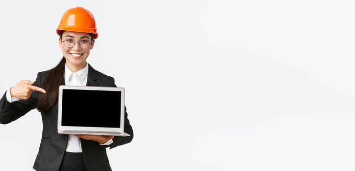 Smiling pleased asian female chief engineer showing chart with enterprise profit, making presentation during investors meeting, wearing safety helmet, pointing at laptop screen, white background