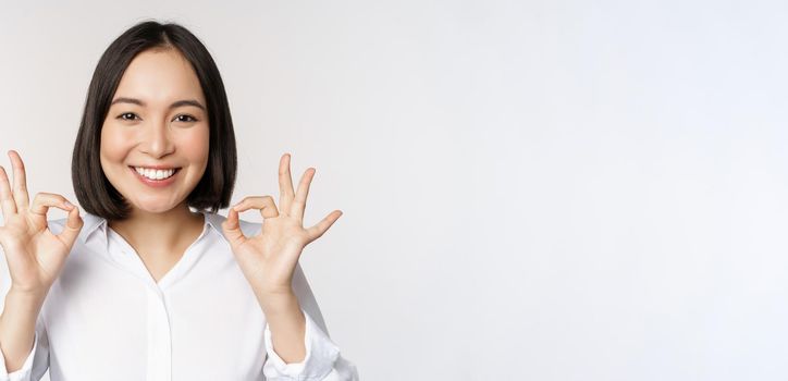 Close up head portrait of asian girl showing okay, ok sign and smiling satisfied, recommending, being pleased, praise and make compliment, white background