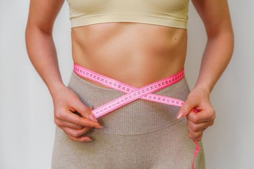 Cropped view of slim woman measuring waist with tape measure at home, close up. An unrecognizable European woman checks the result of a weight loss diet or liposuction indoors. Healthy lifestyle.