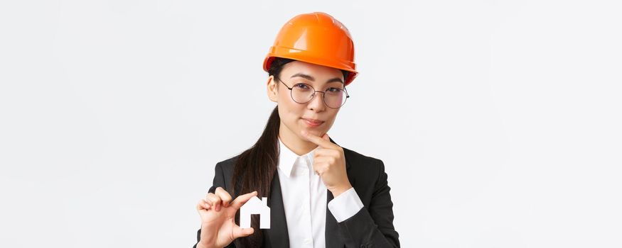 Thoughtful creative female asian architect in helmet and business suit creating new ideas of design, showing home maket and thinking, making cunning face, pondering plan of construction