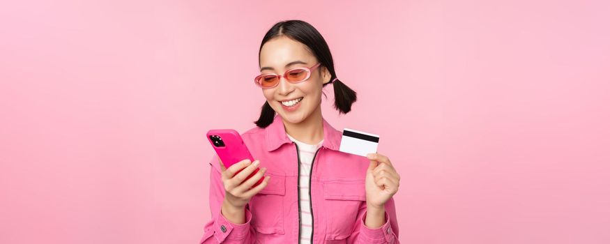 Online shopping. Smiling asian girl shopper, holding smartphone and credit card, paying in mobile app, standing over pink background