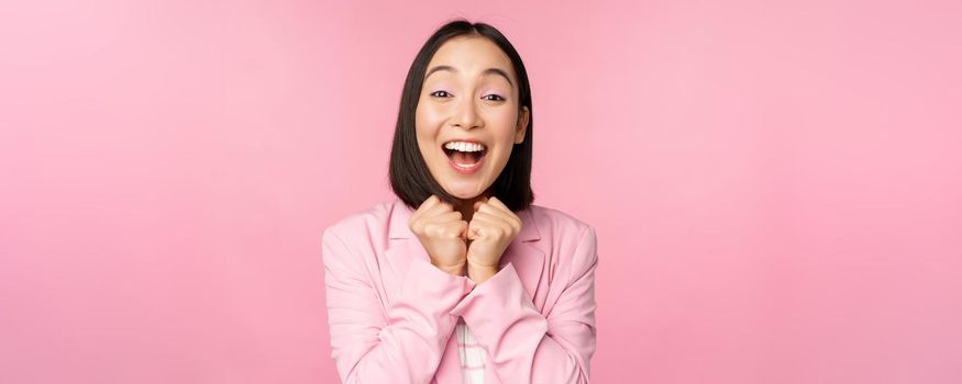 Portrait of happy asian office lady, ceo businesswoman in suit rejoicing and laughing, winning, celebrating, achieve goal and rejoice, standing over pink background