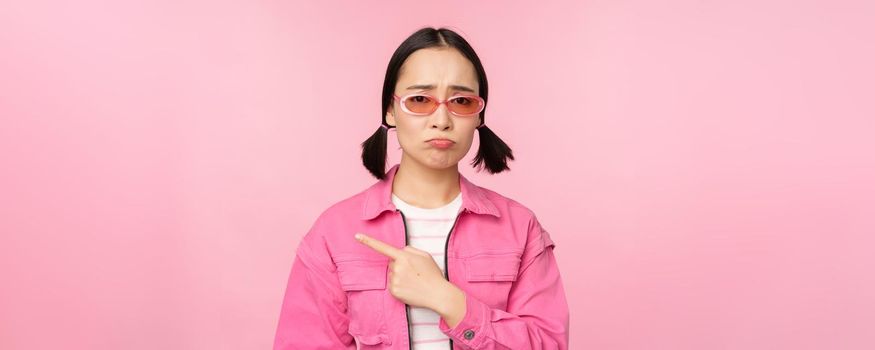 Potrait of cute korean girl in sunglasses, pointing left and looking disappointed, sulking upset, standing over pink background