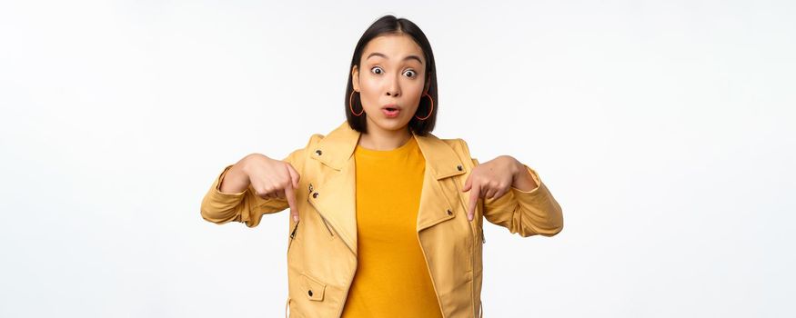 Portrait of surprised asian girl, looking amazed and impressed, pointing fingers down, showing banner with shocked face expression, white background