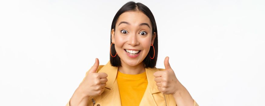 Smiling asian girl showing thumbs up, looking pleased, approve smth, standing over white background