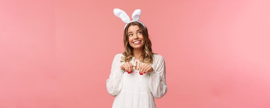 Holidays, spring and party concept. Cute romantic young blond girl imitating bunny, wear white dress lovely rabbit ears, make hand-paws near chest and look dreamy up with daydreaming smile